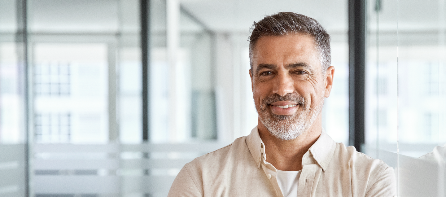 Foto de um homem com cabelos curtos e grisalhos, barba branca e rala, sorrindo. Ele tem idade por volta dos 50 anos e veste uma blusa bege claro por cima de uma camiseta branca. Atrás dele, vemos paredes divisórias de vidro, sugerindo que ele está no seu ambiente de trabalho. Do lado esquerdo da foto, há o texto “Planos Odontológicos Empresariais. O melhor negócio para empresas com mais de 99 funcionários.