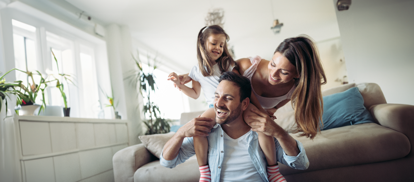 Família feliz em momento de diversão em casa. Pai carregando a filha pequena nos ombros enquanto a mãe os abraça, todos sorrindo e vestindo roupas claras em um ambiente aconchegante de sala de estar.