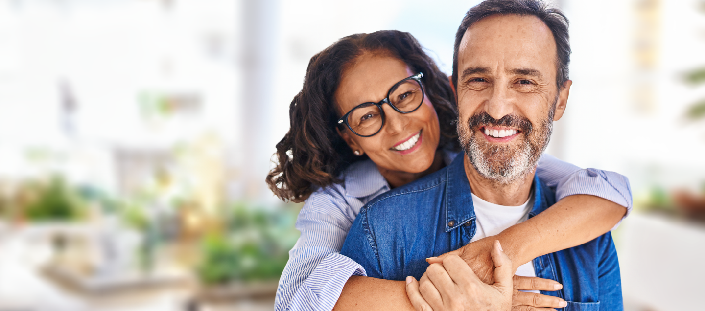 Foto de um casal abraçado sorrindo. Os dois têm idade por volta de 50 anos. A mulher é branca, tem cabelos escuros, usa óculos e uma camisa listrada de azul e branco. O homem é branco, tem cabelos grisalhos, barba branca e veste uma camisa jeans por cima de uma camiseta branca. Do lado esquerdo da foto, há o texto “Plano Sigma. O básico pra fazer você feliz. Apenas R$ 39,90 por mês.